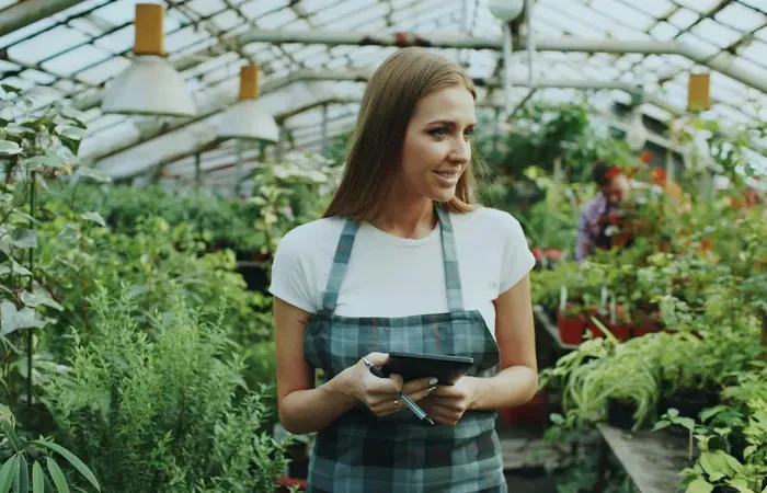 Smiling greenhouse worker holds a tablet among lush plants and pots, managing listings and customer messages